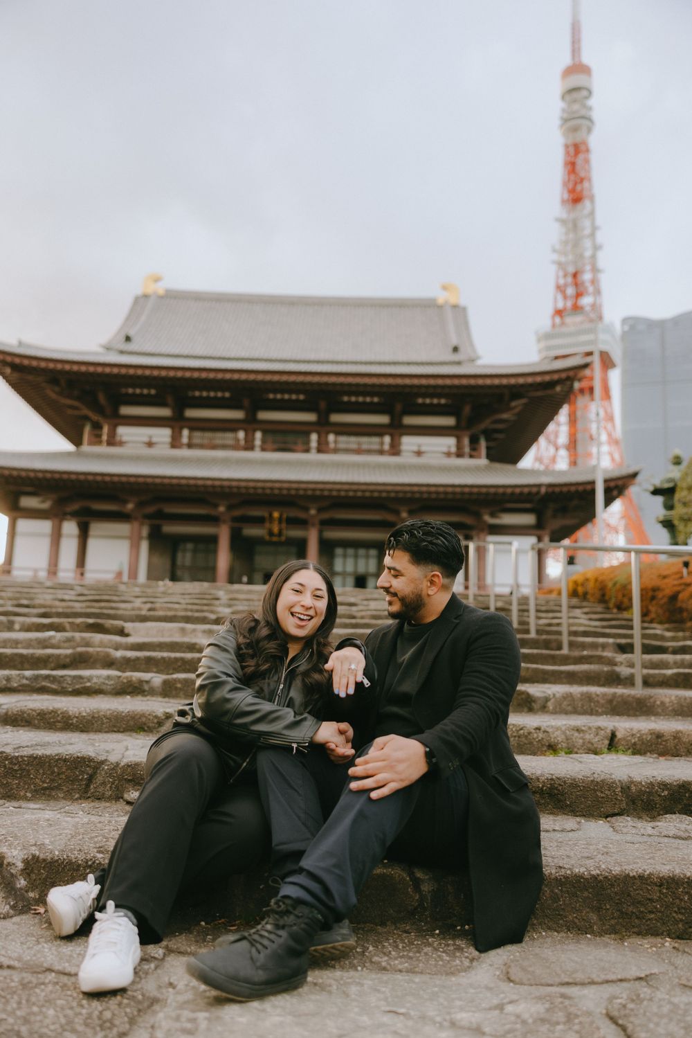 A couple sits on stone steps outside Zojoji Temple with Tokyo Tower visible in the background on a cloudy day in Japan.
