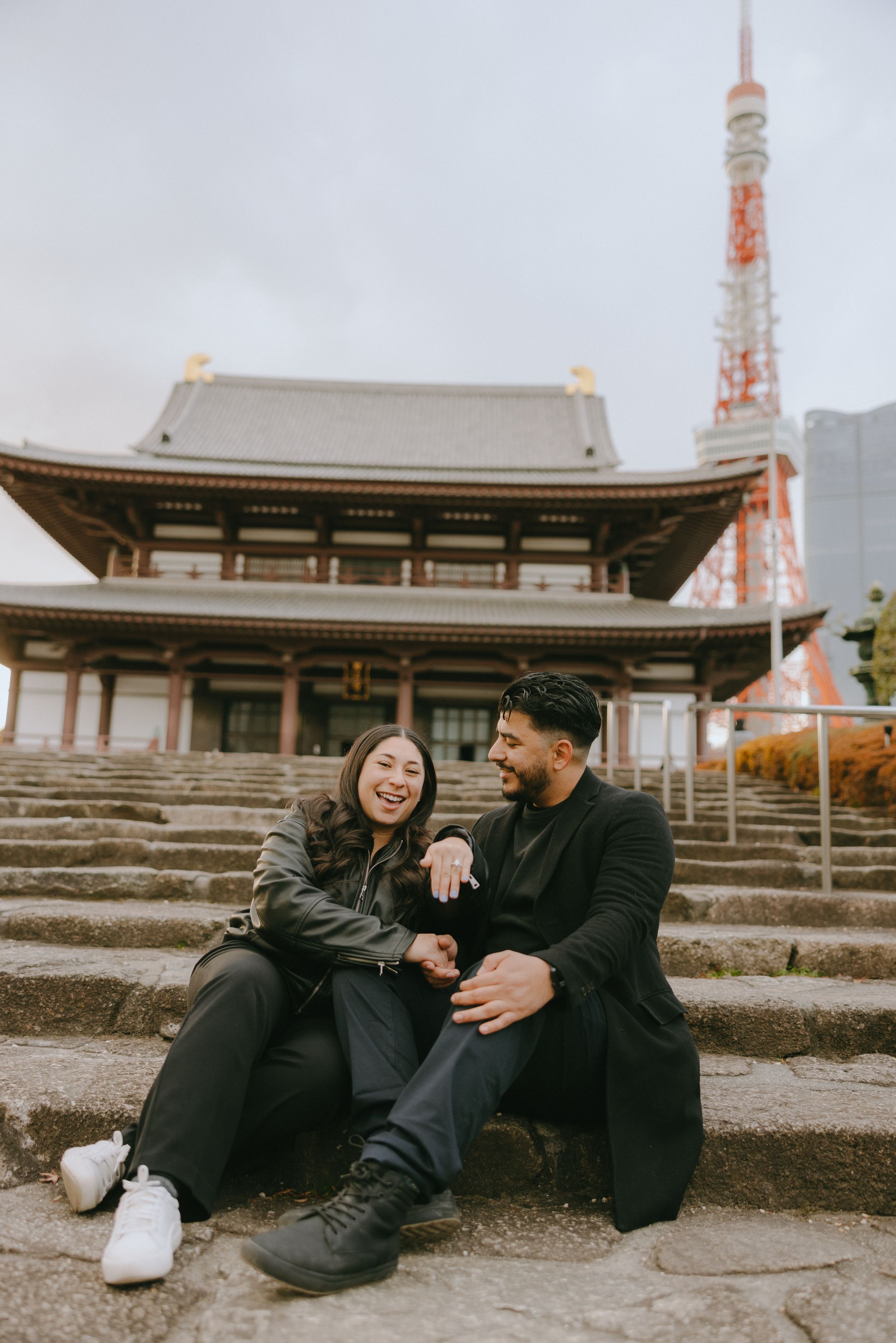A couple sits on stone steps outside Zojoji Temple with Tokyo Tower visible in the background on a cloudy day in Japan.