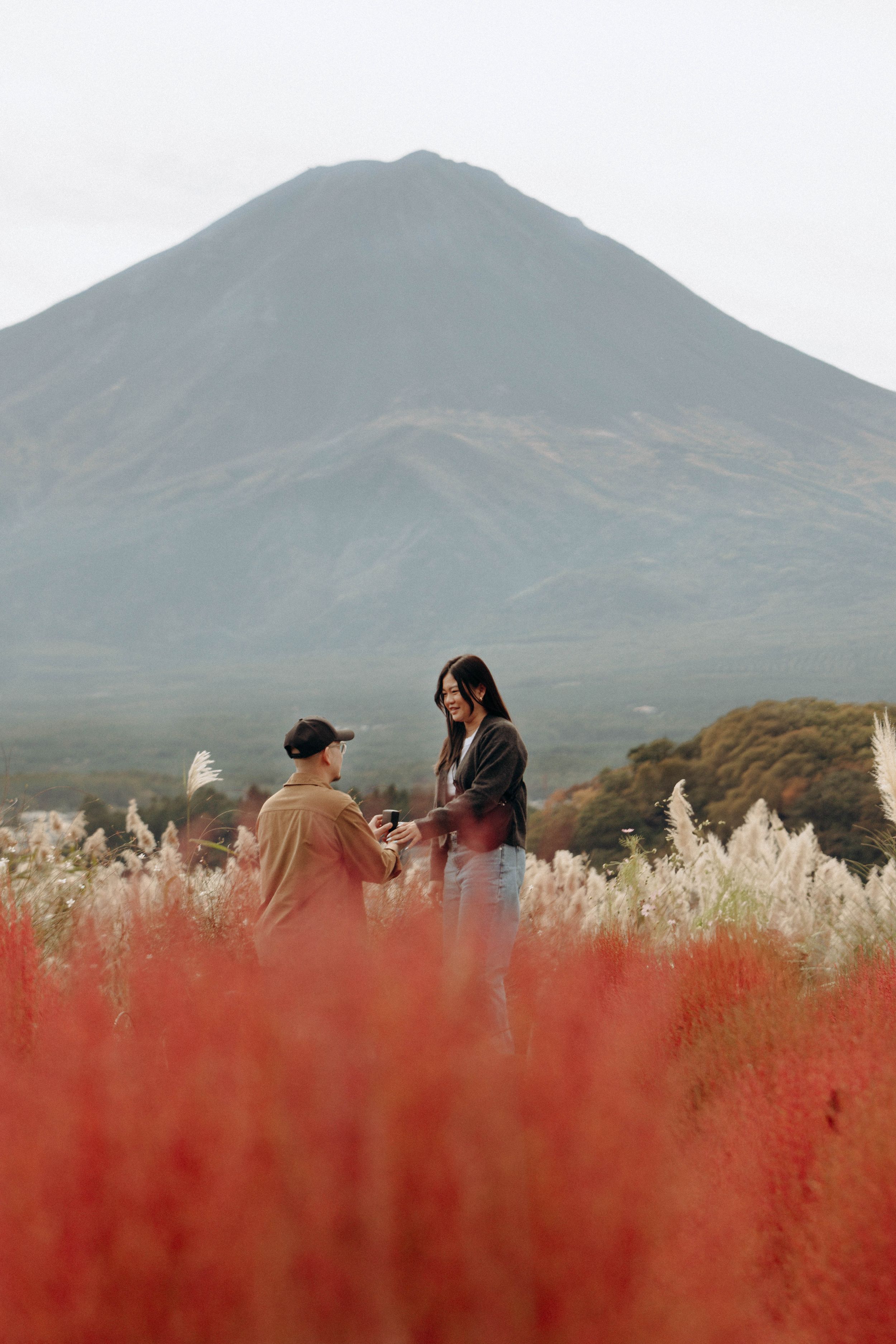 Two people share an intimate moment in a field of red grass with a majestic mountain in the background.