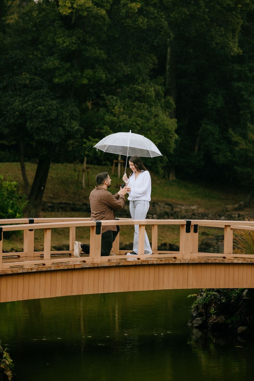 A romantic proposal moment unfolds on a wooden bridge over a pond with a white umbrella in a lush garden setting.