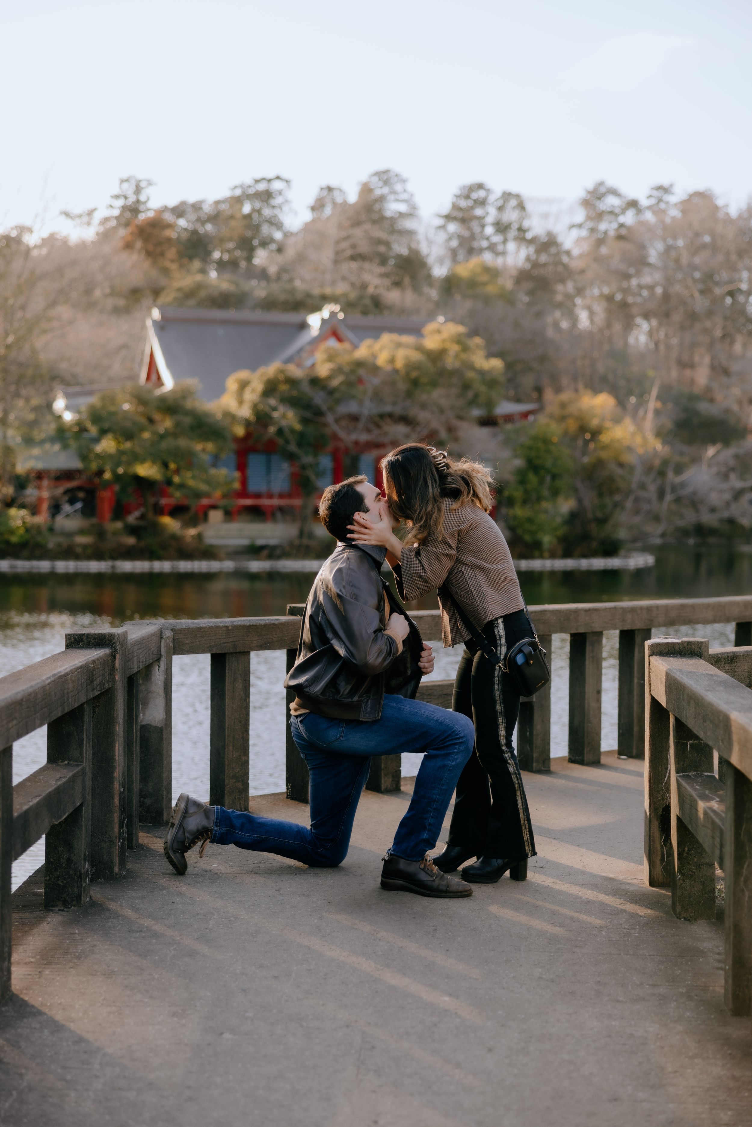 A heartwarming proposal moment unfolds on a wooden bridge at a Japanese garden during sunset.