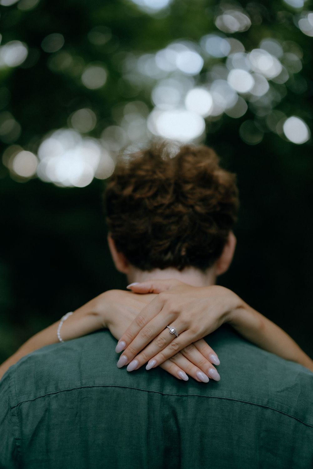 Two hands with a wedding ring crossed behind a green shirt under soft bokeh lighting in nature.