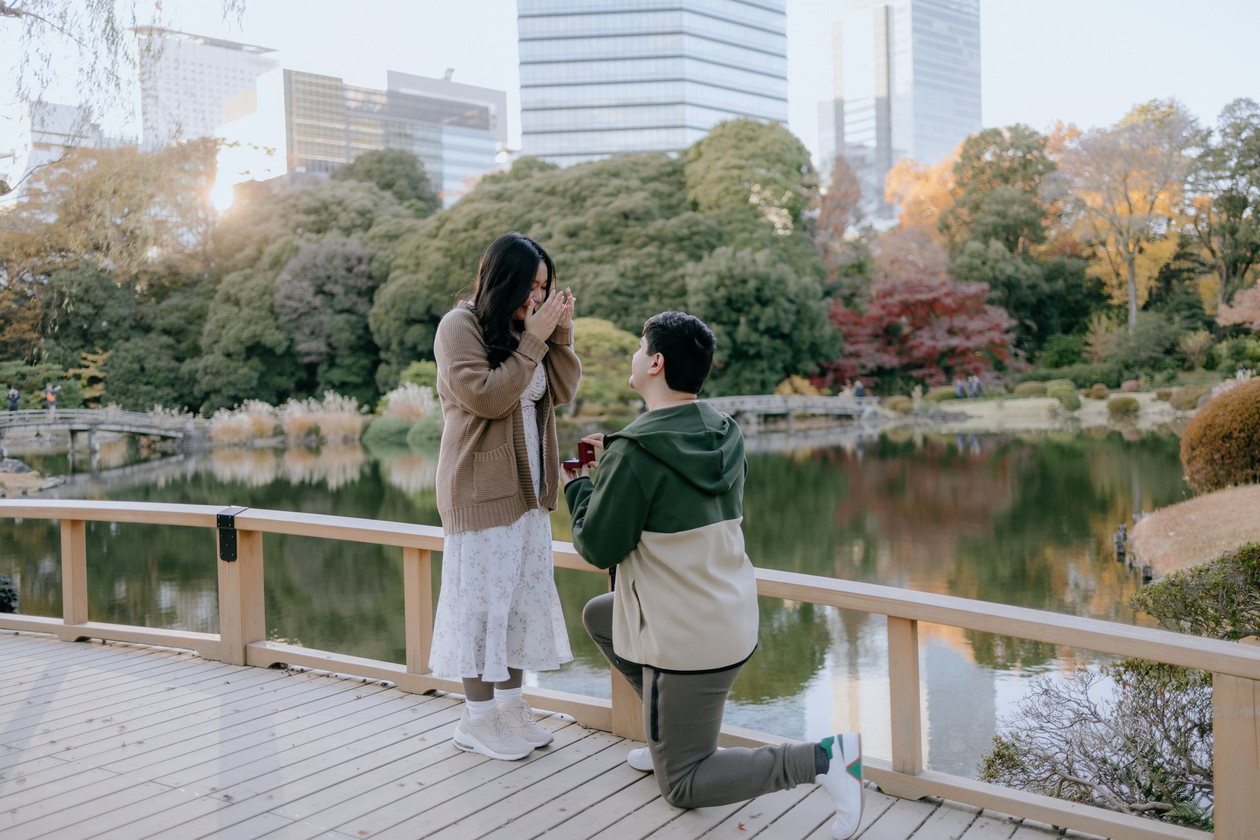 A couple captures photos from a wooden boardwalk overlooking a serene lake in an autumn park with city buildings in background.