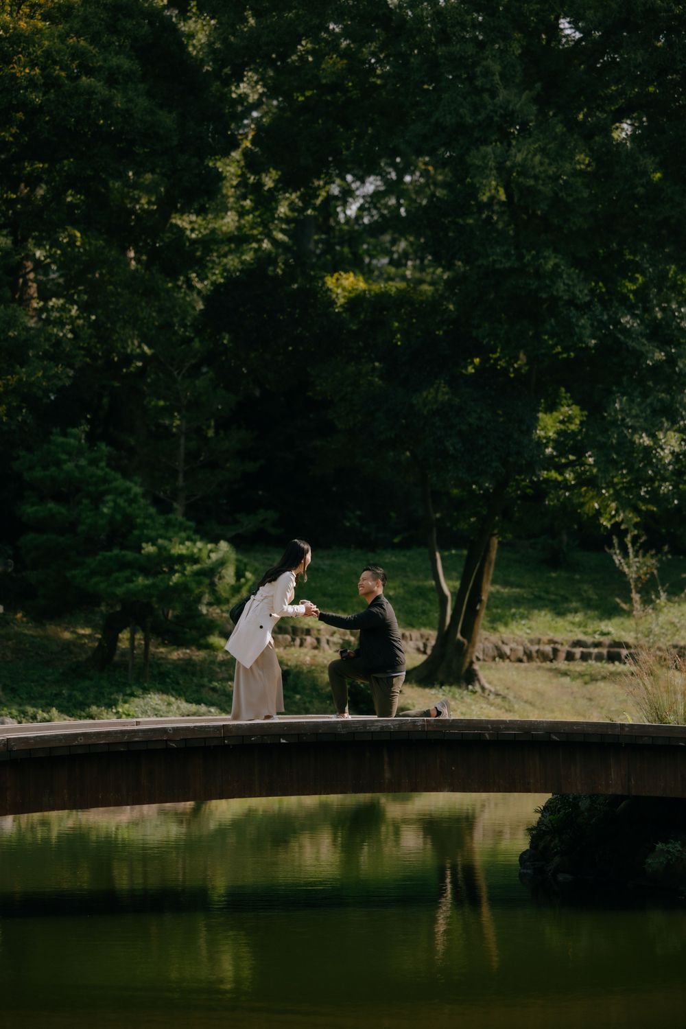 A proposal moment on an arched bridge over a serene pond in a lush garden setting.