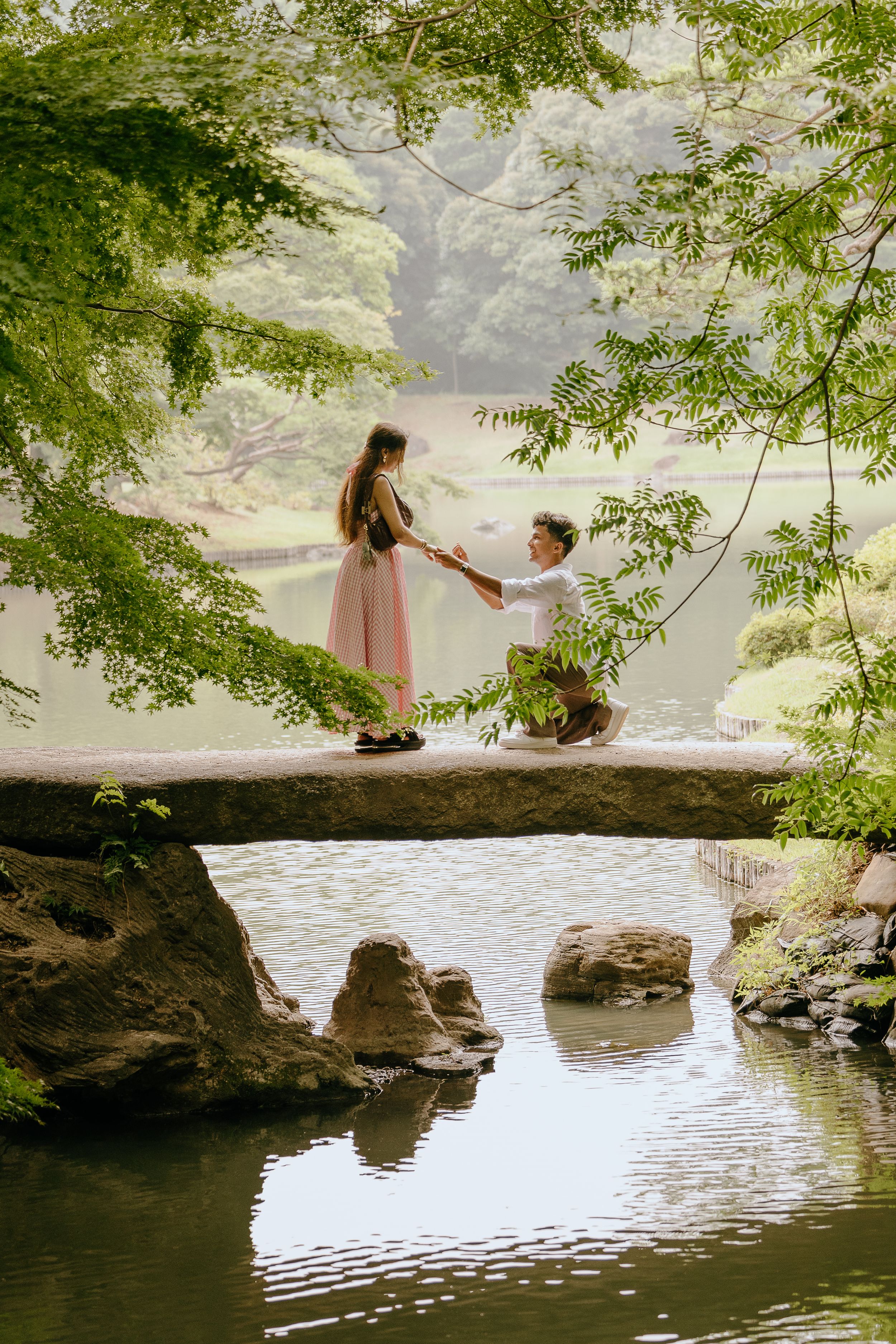 Two people sit together on a large rock formation overlooking a serene lake surrounded by lush greenery.