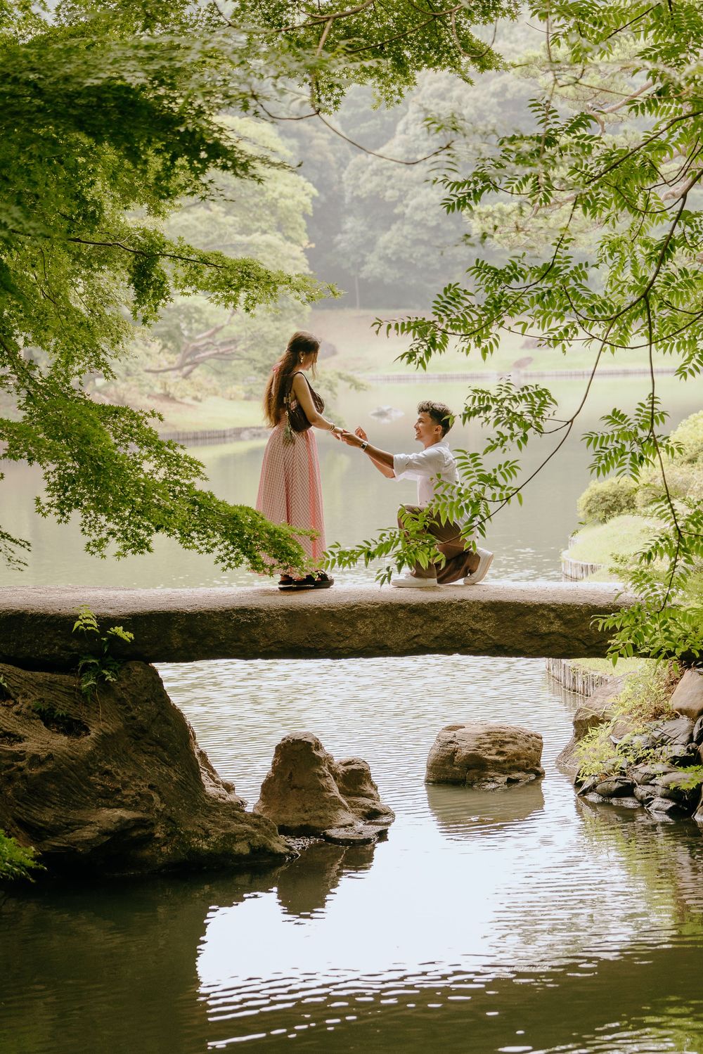 Two people sit together on a large rock formation overlooking a serene lake surrounded by lush greenery.