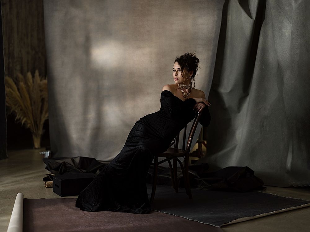 Woman in elegant black strapless gown seated on chair against draped gray backdrop in dramatic studio portrait.
