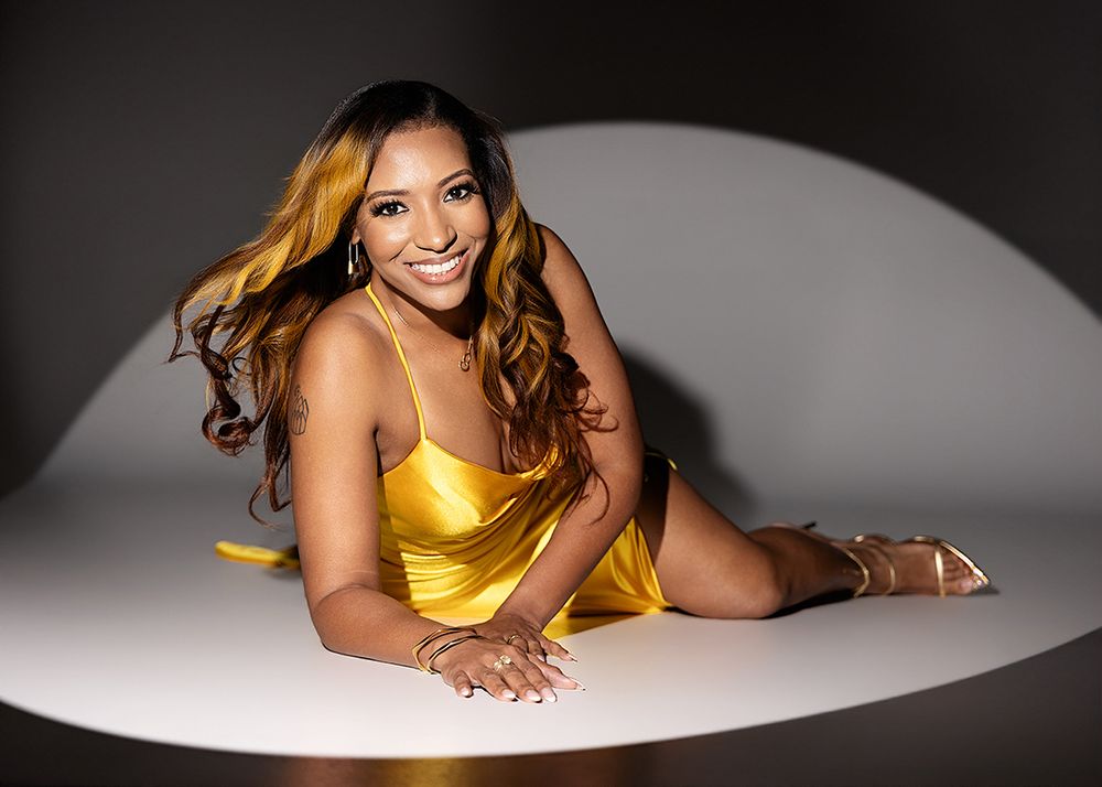 A woman in a golden yellow dress poses gracefully on a white surface, smiling with flowing highlighted hair.