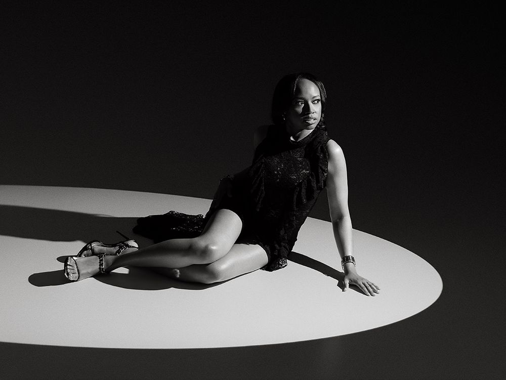 Black and white photo of a woman in a black dress posing gracefully on a circular lit floor in a dark studio.