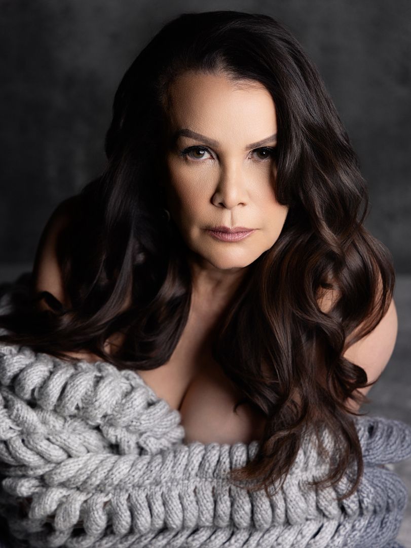 A woman with long dark wavy hair poses in a chunky knit sweater against a dark gray background.