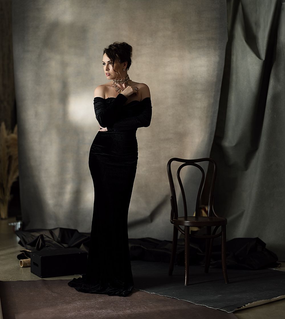 Woman in elegant black off-shoulder gown poses gracefully in a studio with vintage chair and draped backdrop.