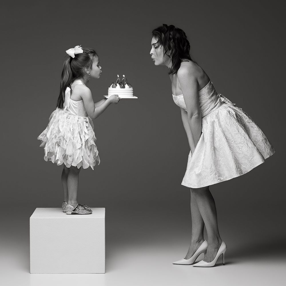 A young girl on a pedestal presents a birthday cake to a woman in a white dress in a black and white photo.