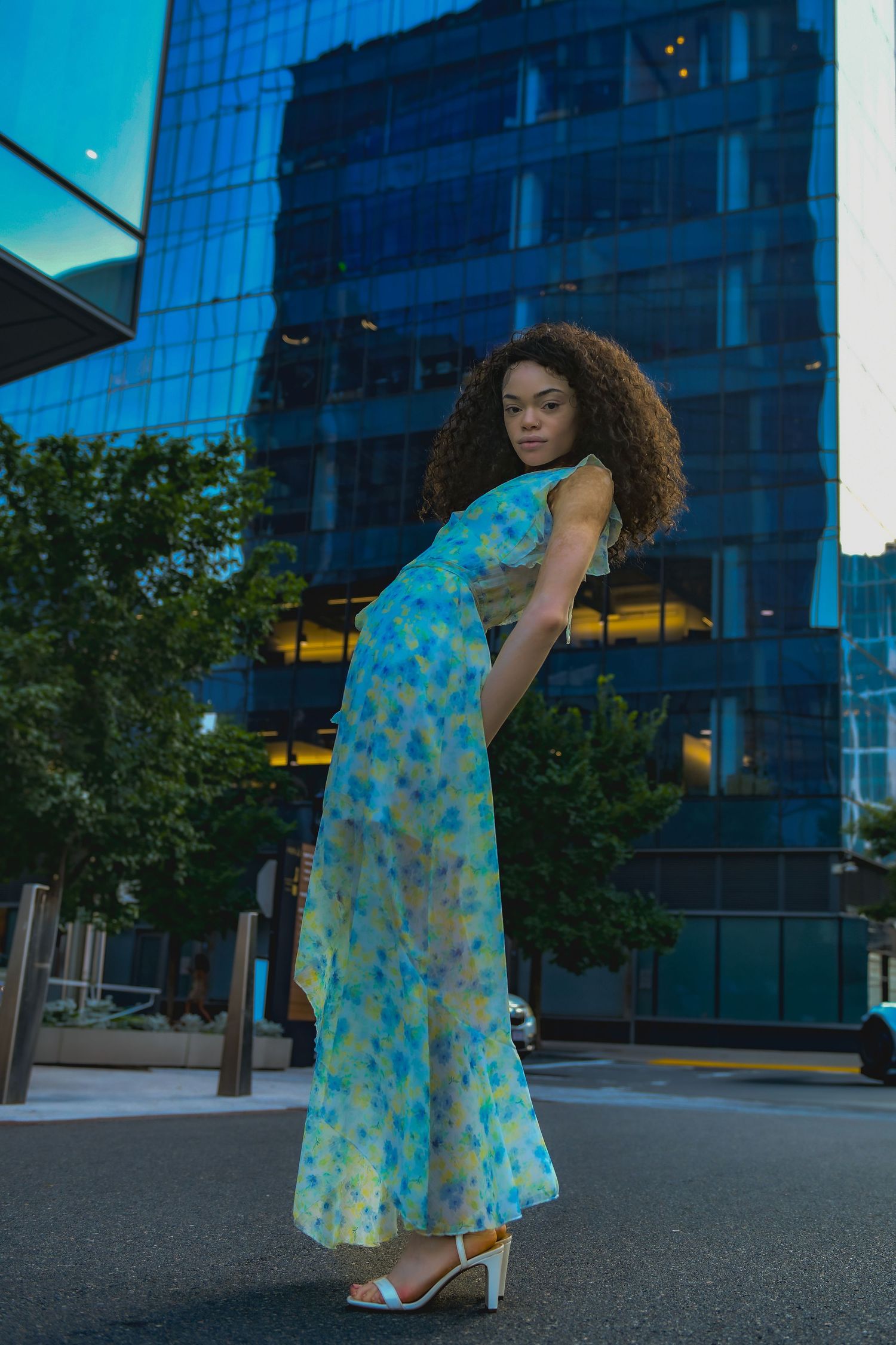 Full length fashion portrait in a flowing floral dress against modern city buildings.