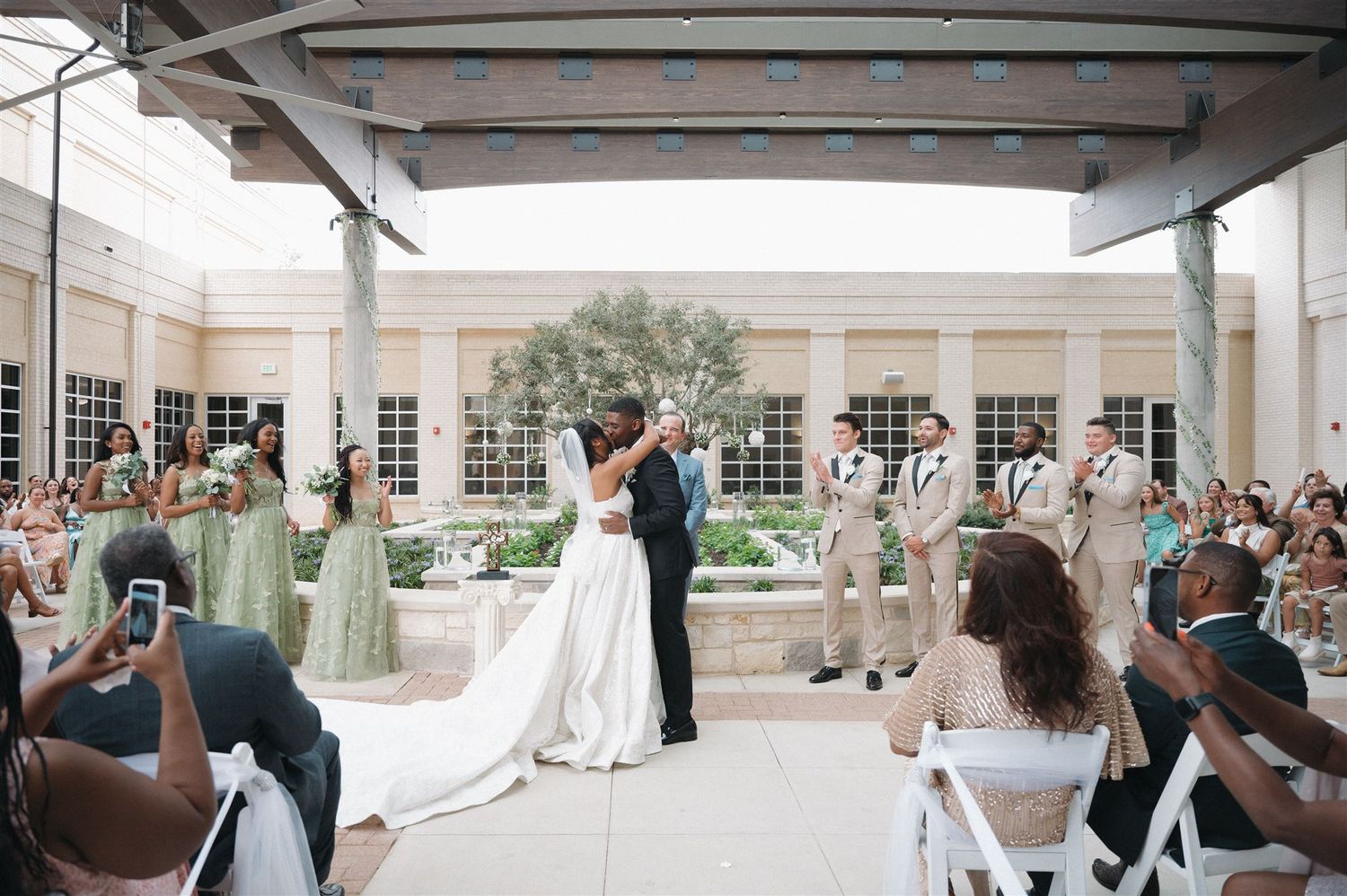 A couple shares their first kiss at an outdoor wedding ceremony while guests capture the moment on their phones.