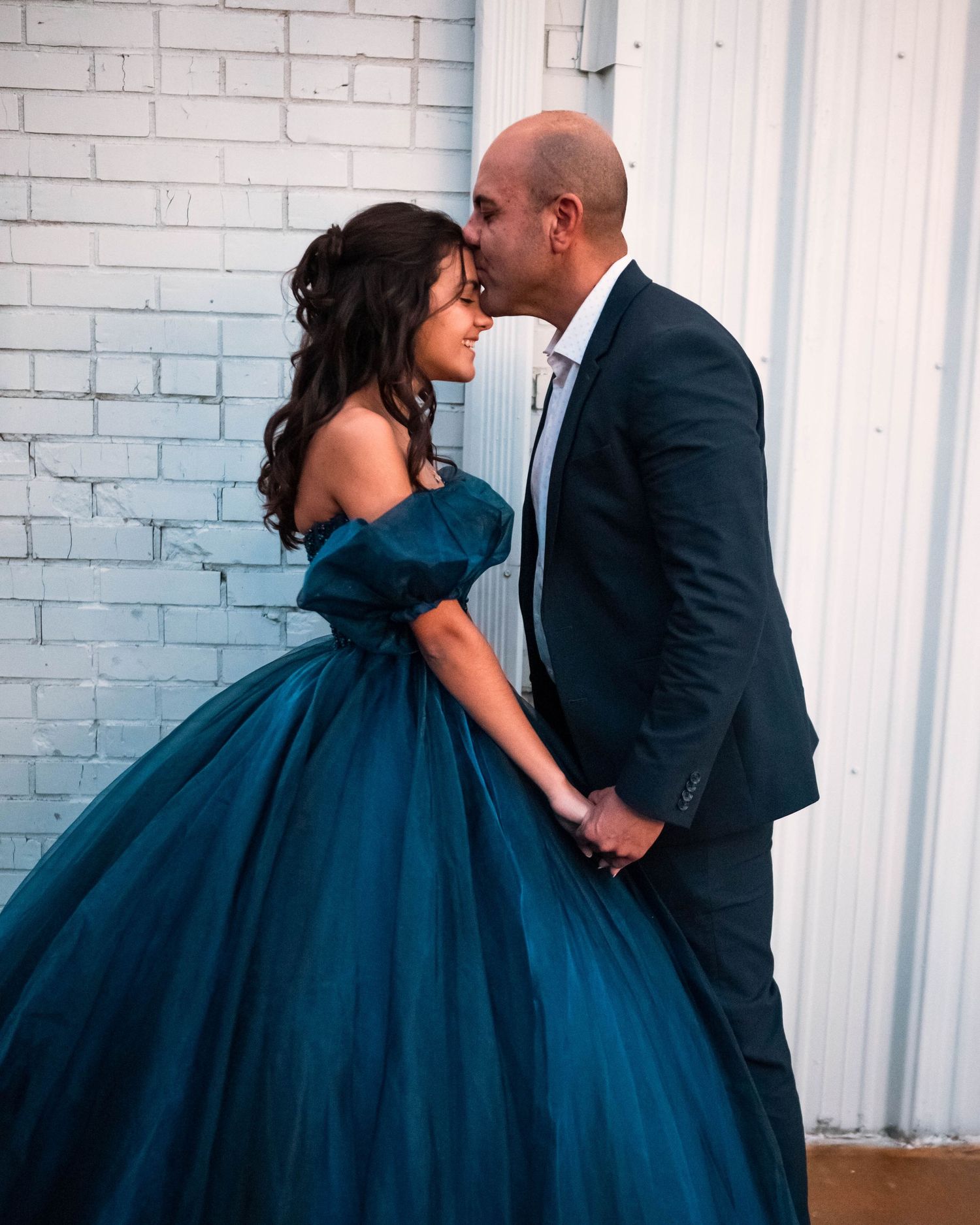 A couple in formal attire poses together against a white brick wall, with her wearing a stunning navy blue ballgown.