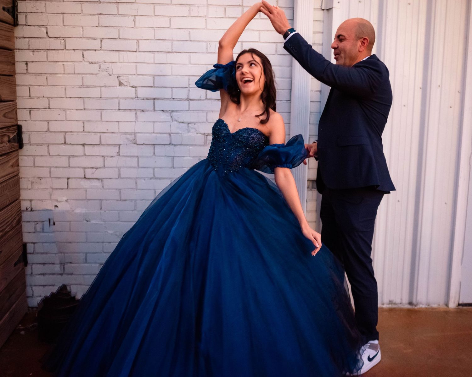 Playful dance poses captured in sequence showing a navy ballgown twirling against white brick backdrop.