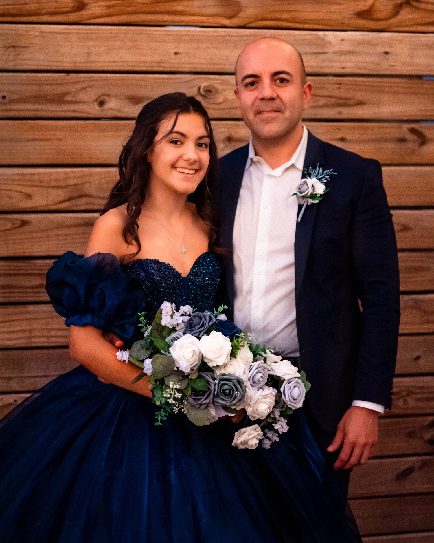 Formal couple poses with white floral bouquet against rustic wooden wall background.