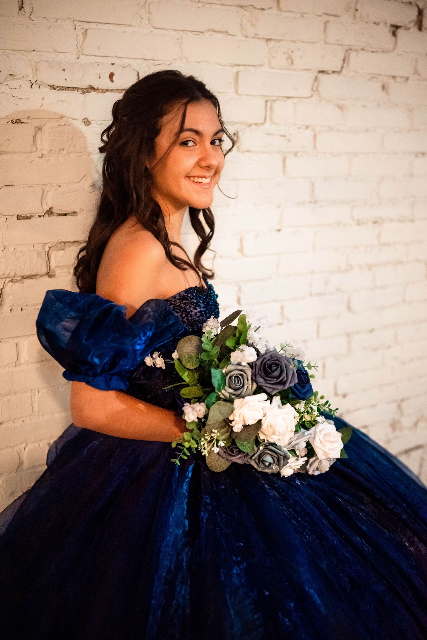 Woman in navy blue ball gown poses with floral bouquet against white brick wall.
