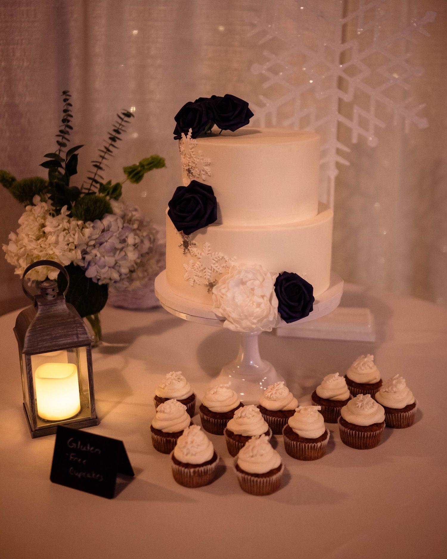 Wedding dessert table with white cake, cupcakes and lantern decorations in warm lighting.