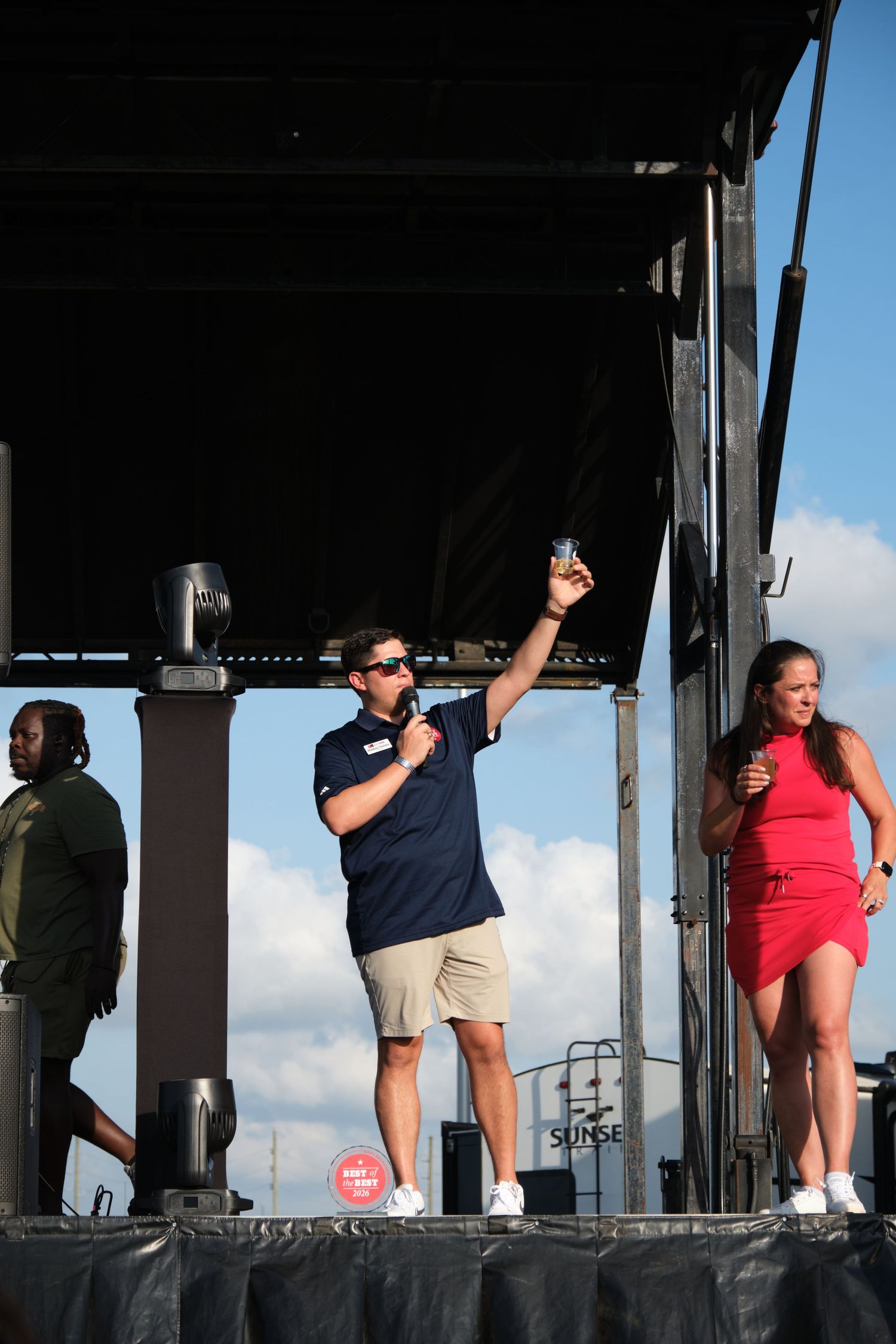 A man in navy polo raises a drink on an outdoor stage while a woman in red dress stands nearby.