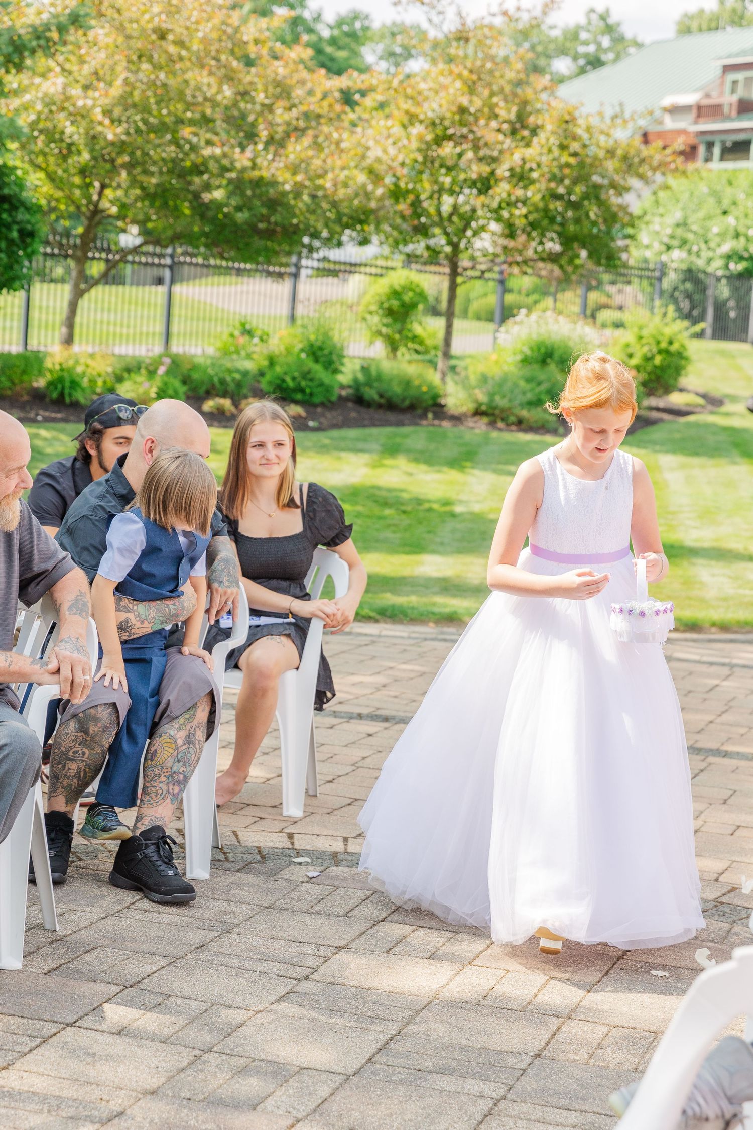 Wedding ceremony guests seated on white chairs on a brick patio with greenery in background.