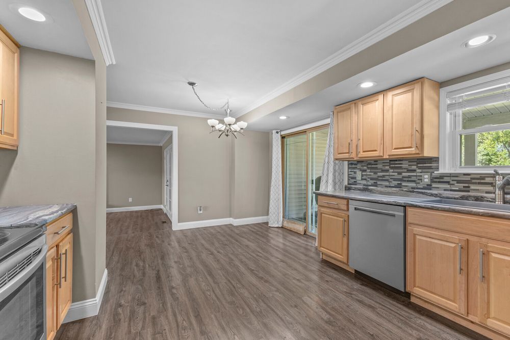 Bright kitchen with light wood cabinets, stainless dishwasher, tile backsplash, and chandelier dining area.