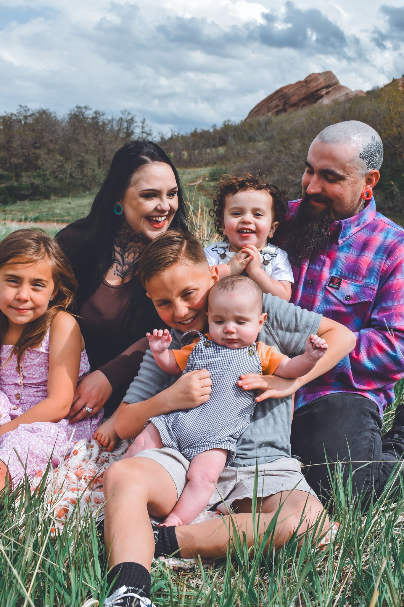 Happy family of six sitting together in green grass with rocky landscape in background.