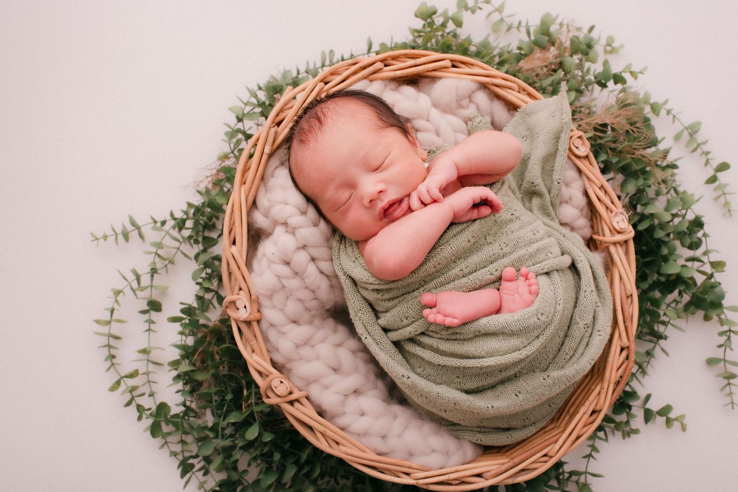 Sleeping newborn baby wrapped in sage green swaddle lying in wicker basket with eucalyptus.
