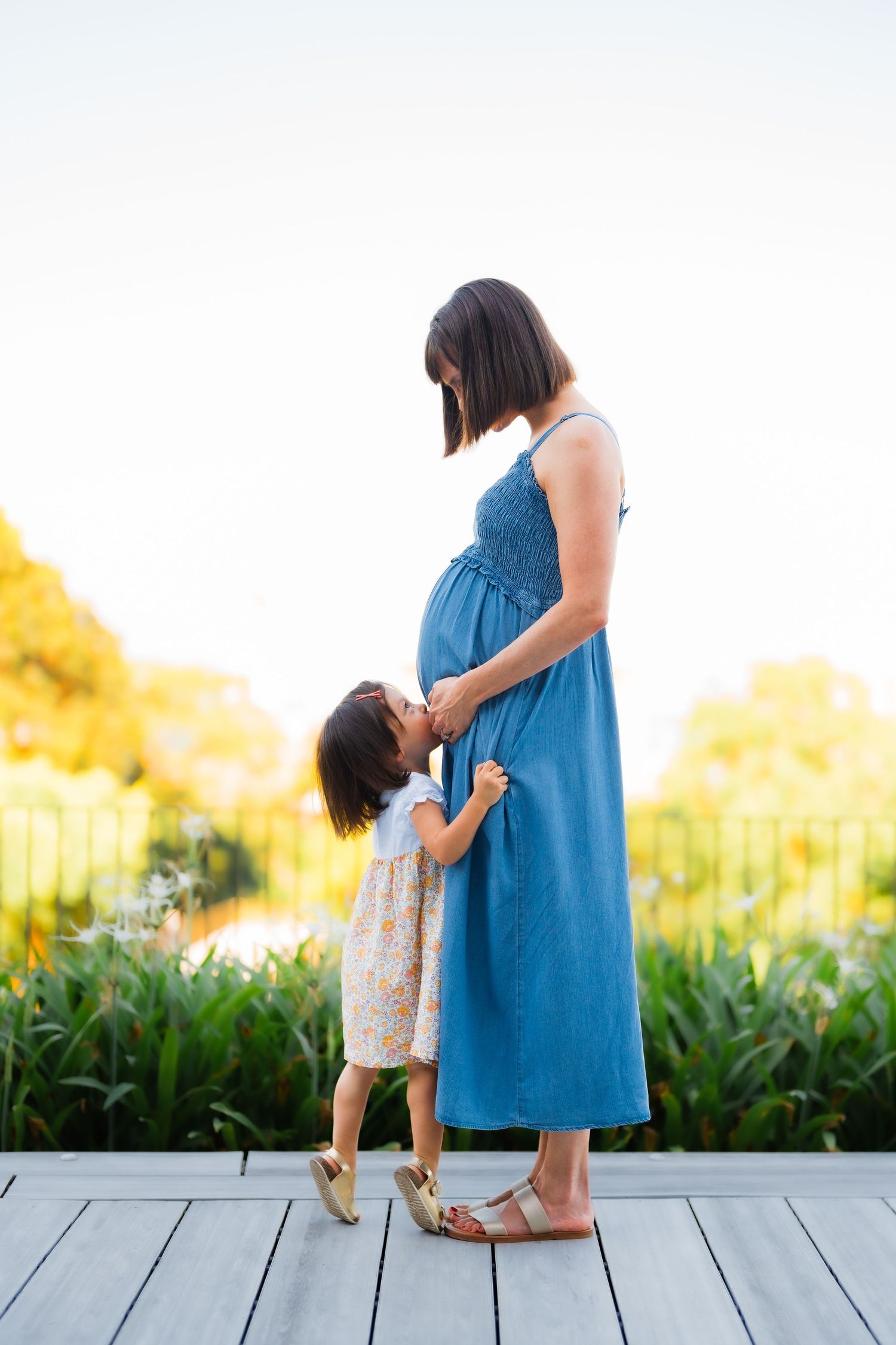 A mother in blue dress stands with small child on wooden deck against golden sunset.