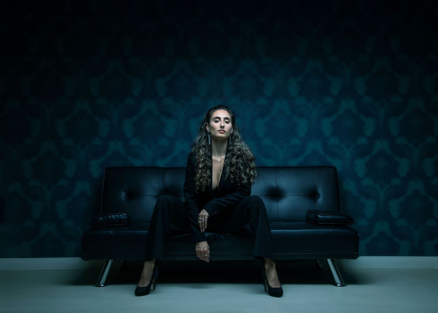 A dramatic portrait against a turquoise damask wall shows someone seated on a black leather couch in dark attire.