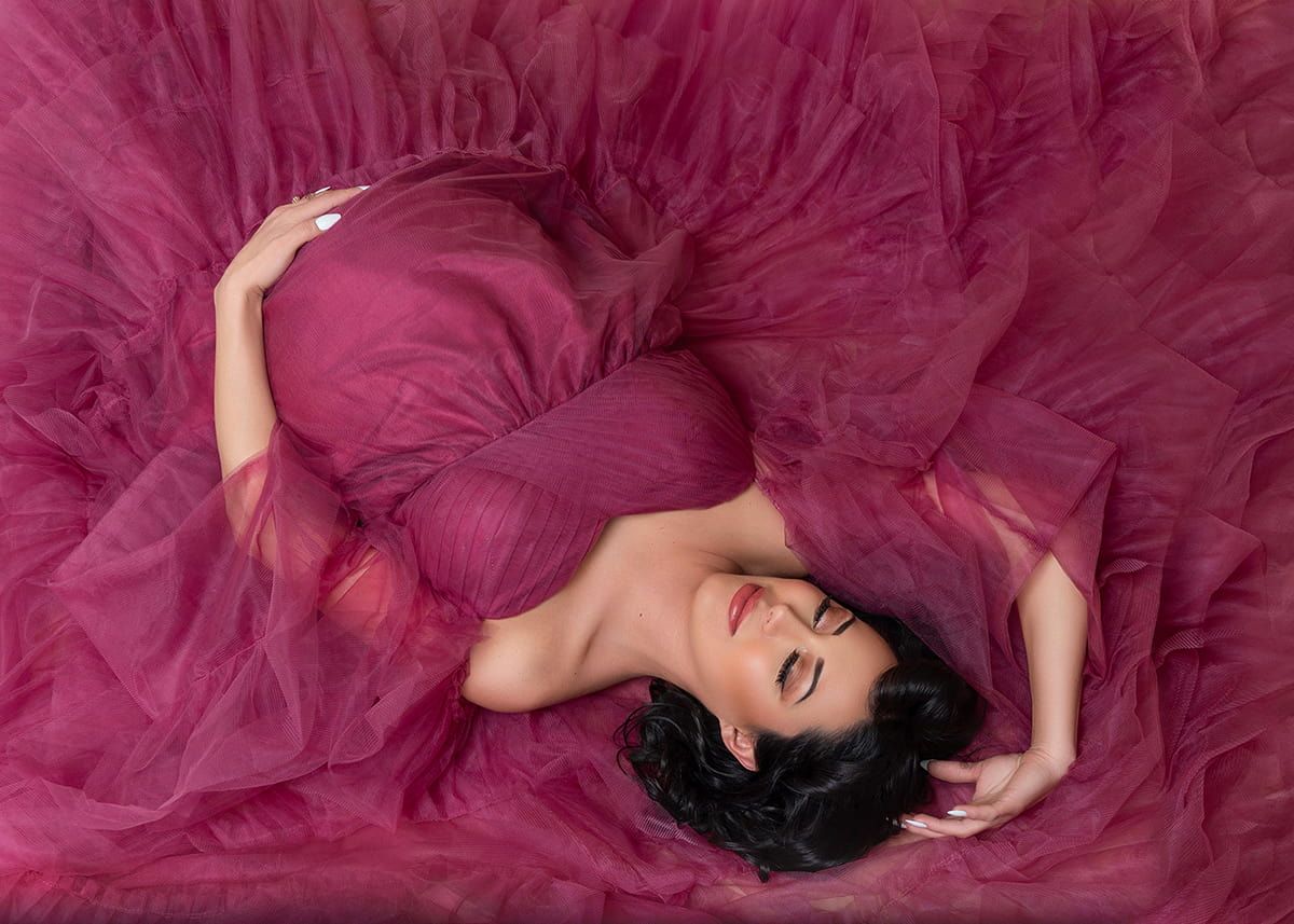 Pregnant woman in flowing burgundy tulle gown lying gracefully in studio, photographed by luxury maternity photographer in Denver.