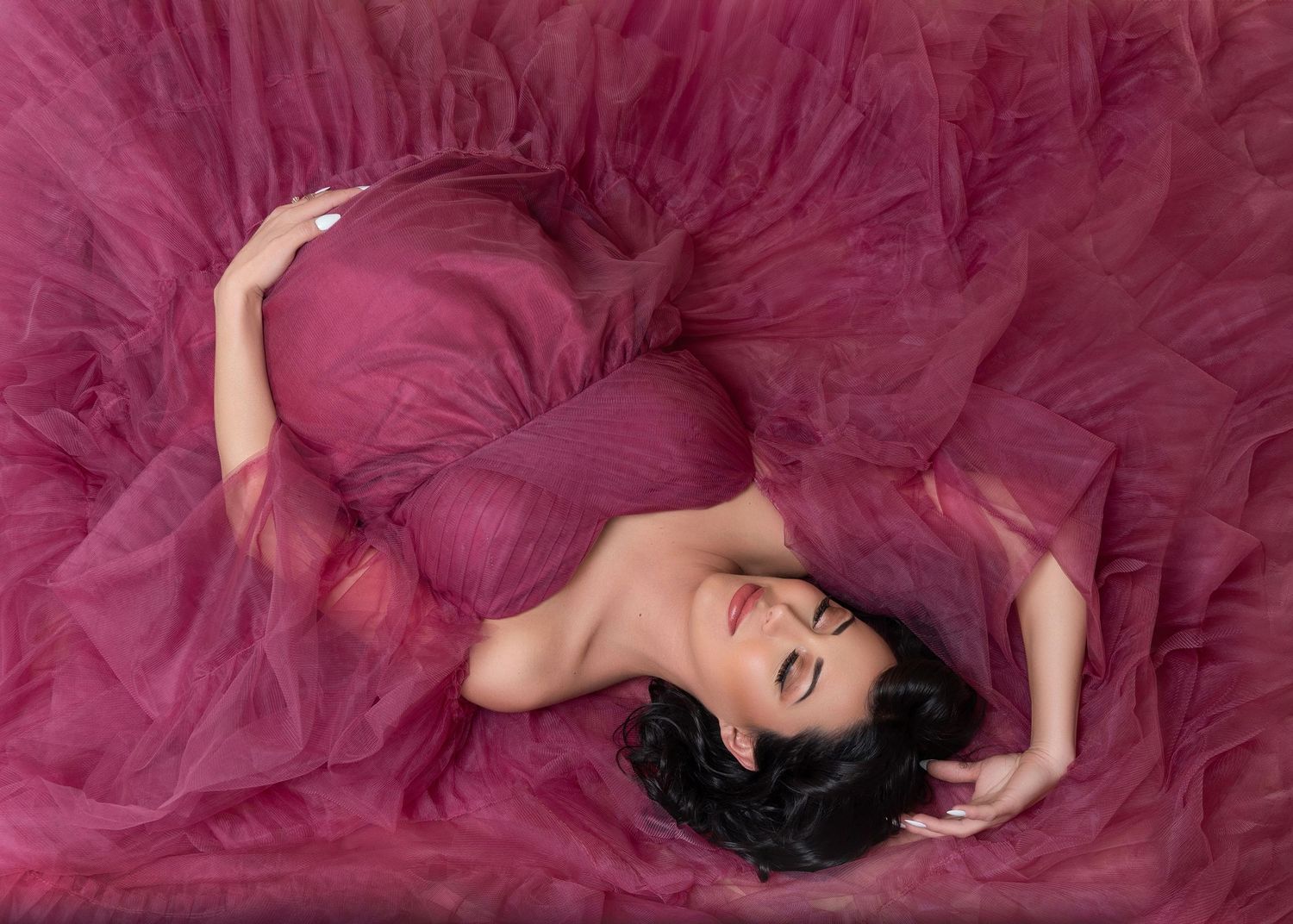 A pregnant mother in a flowing magenta tulle gown lays gracefully on the floor during an artistic maternity portrait session.