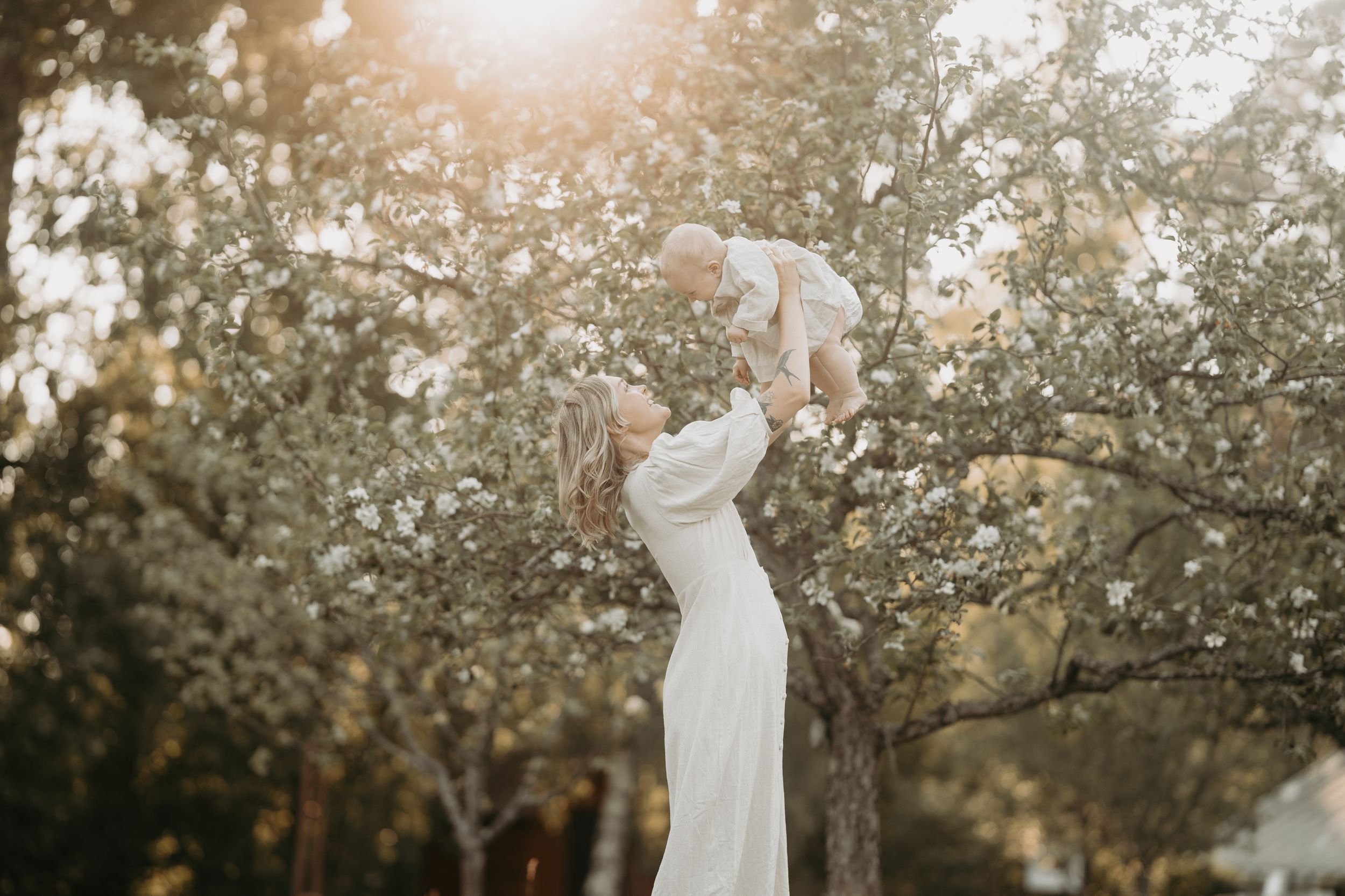 Joyful moment captured in sunlit garden with flowing white dress and baby lifted high.