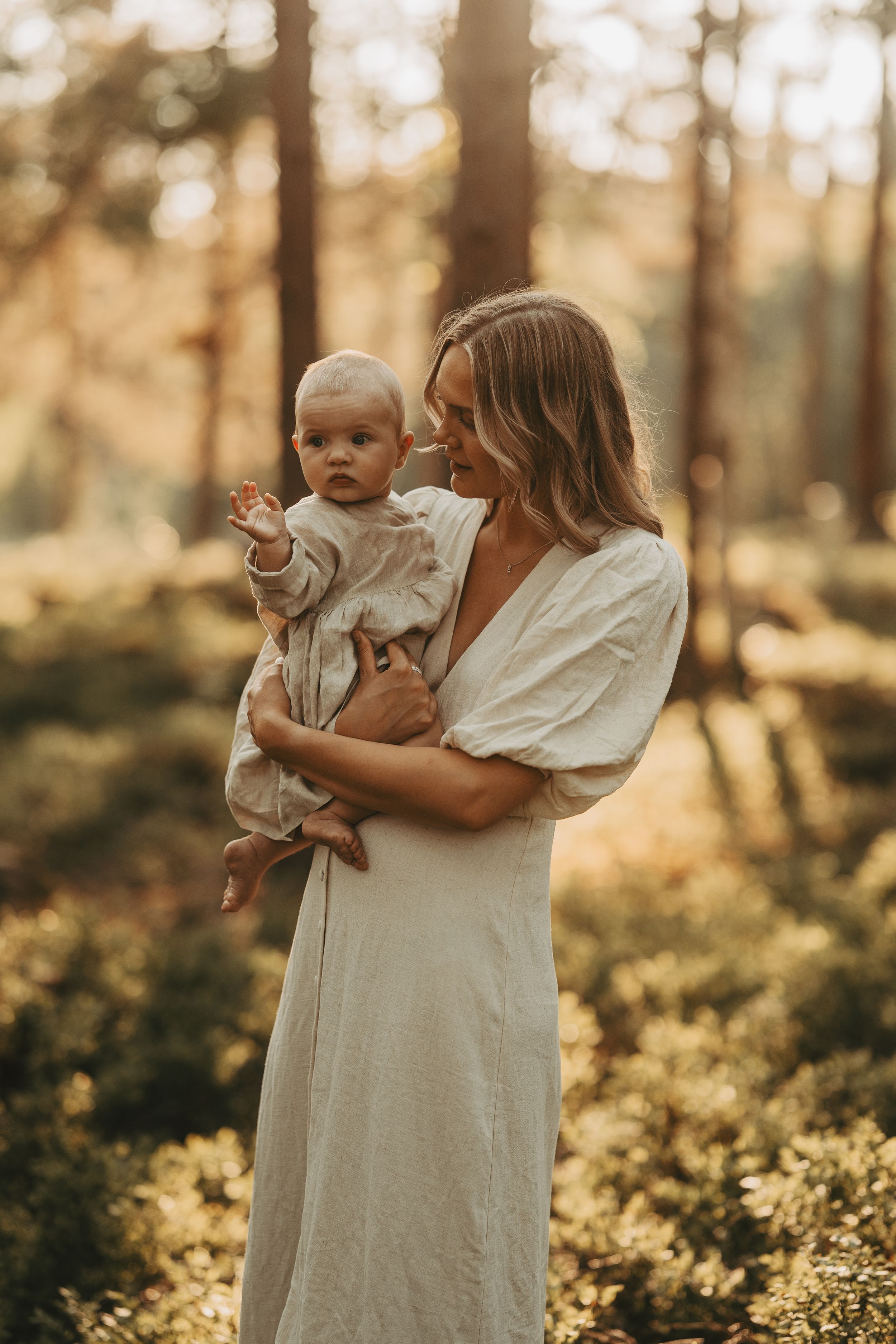 Parent and infant in white clothing share tender moment in sunlit forest setting.
