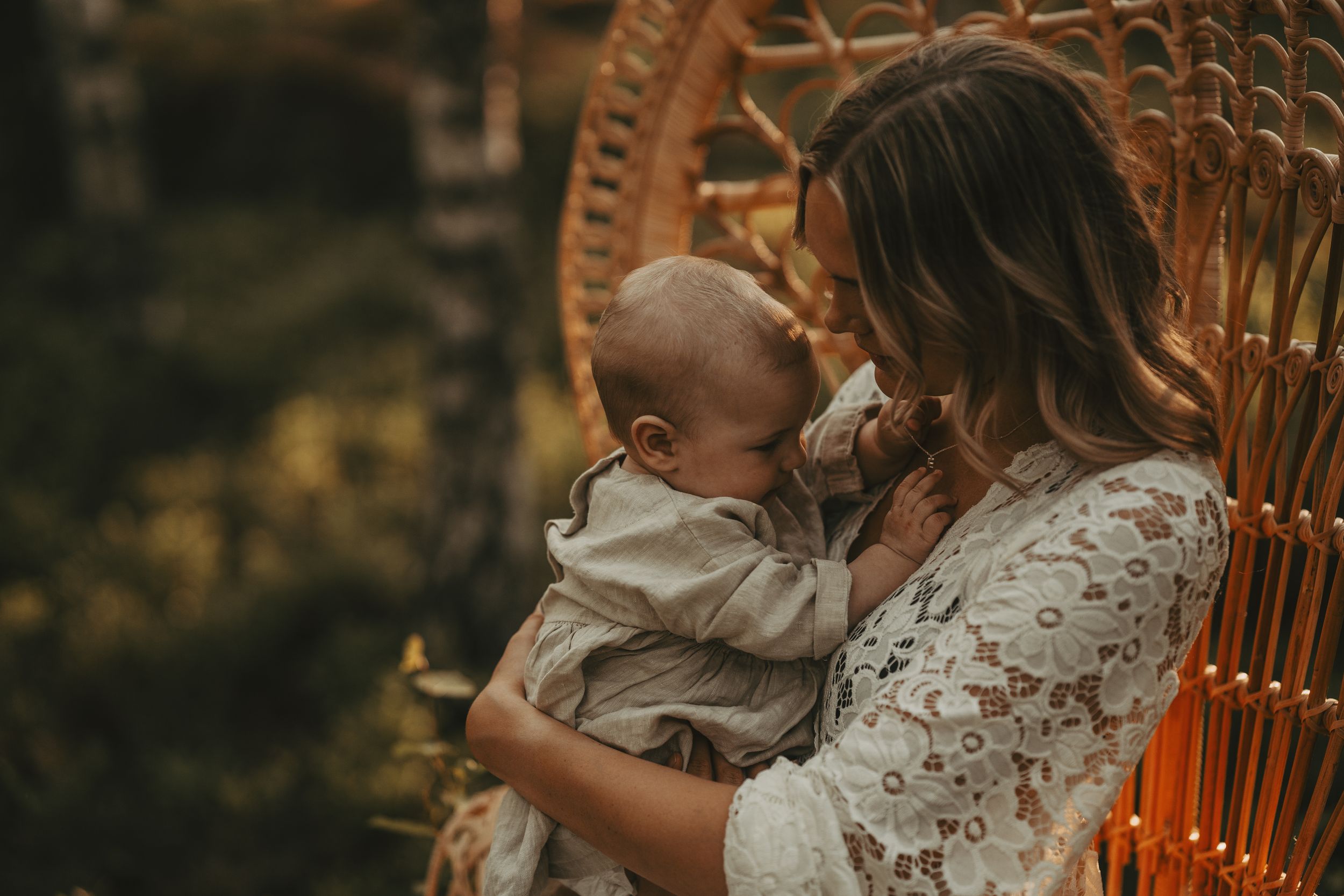 Parent and baby sharing intimate moment in wicker chair during outdoor photo session.