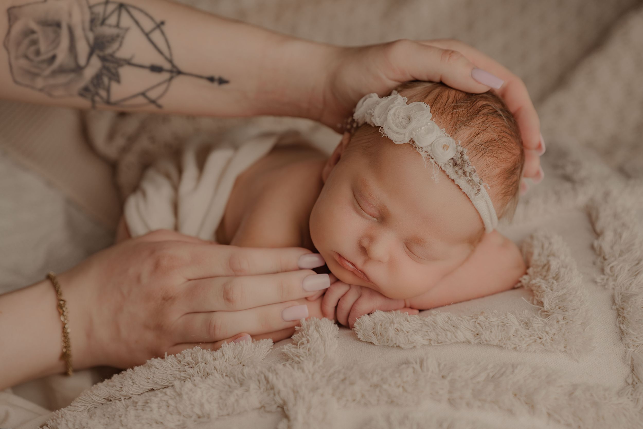Newborn baby girl sleeping peacefully on fluffy blanket wearing white floral headband while mother gently cradles her head.