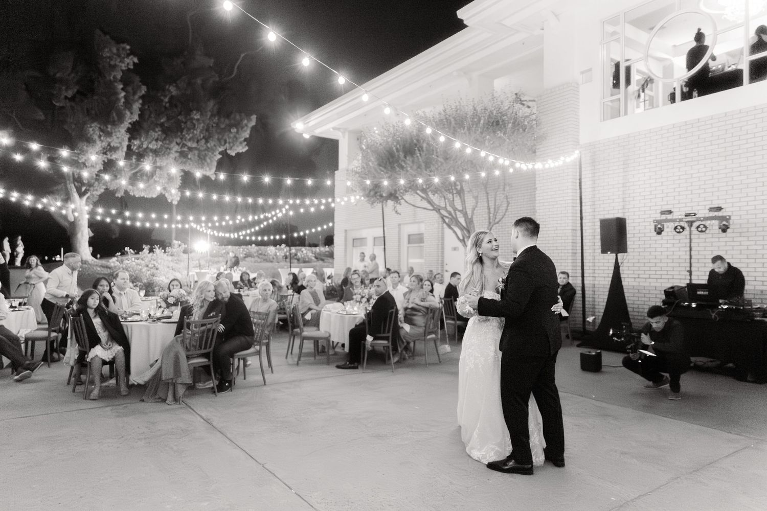 Wedding guests dance under romantic string lights at an outdoor evening reception with tables and decor visible.