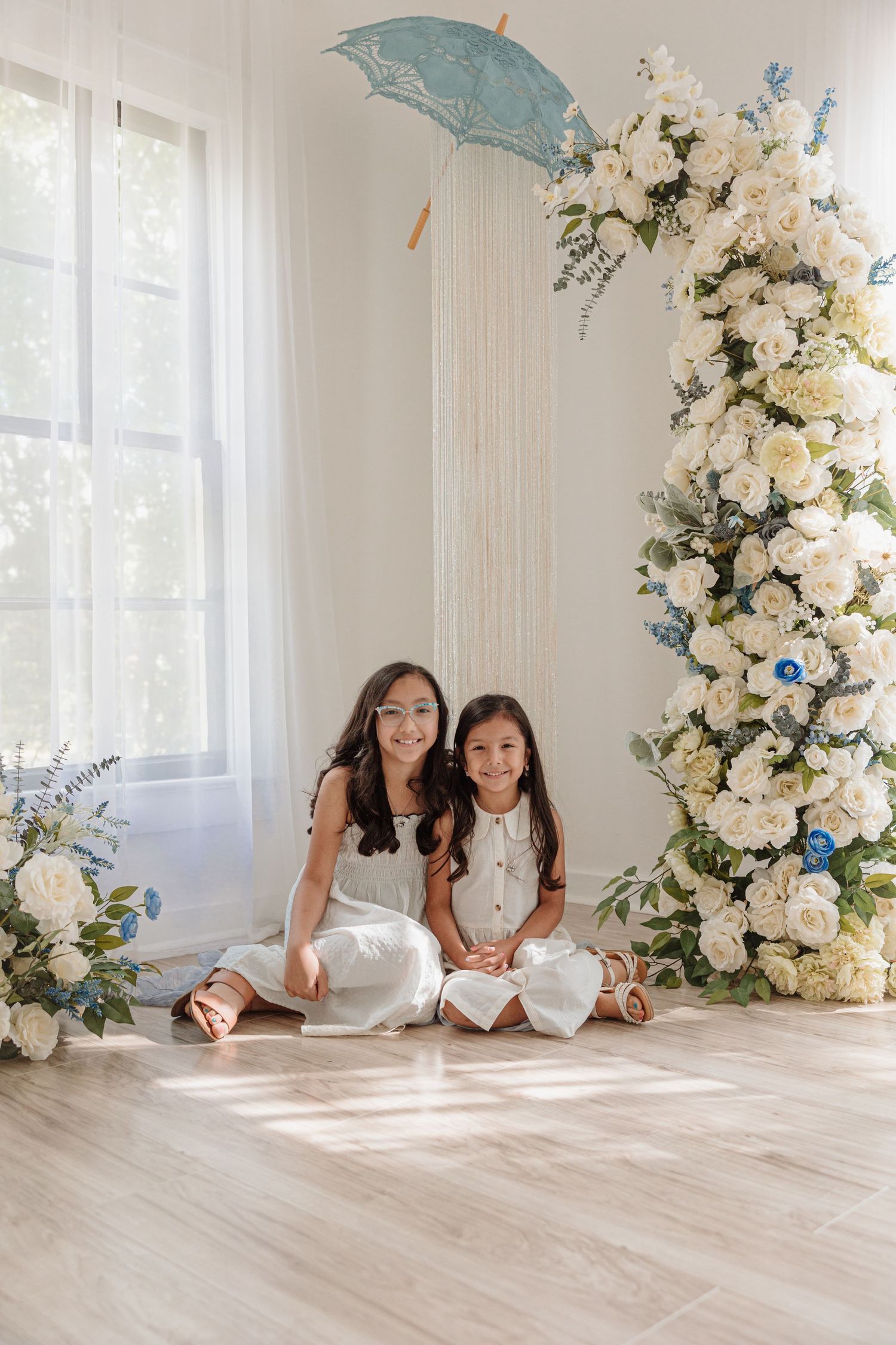 Indoor portrait setting with lace parasols and elegant white floral arrangements against a light wall.
