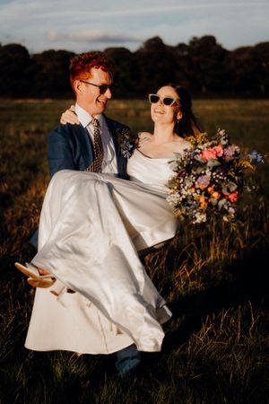 A romantic wedding photo shows a groom lifting his bride in a white dress as they laugh together in a sunset field.