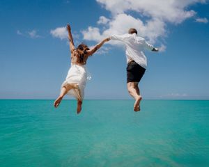 Joyful couple in white attire jumping together over crystal clear turquoise waters.
