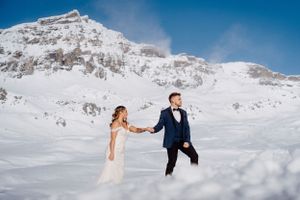 Newlyweds walking through snowy mountain landscape in winter wedding attire.