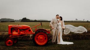 Elegant wedding couple poses beside a vintage bright red tractor in a rural countryside setting at sunset.