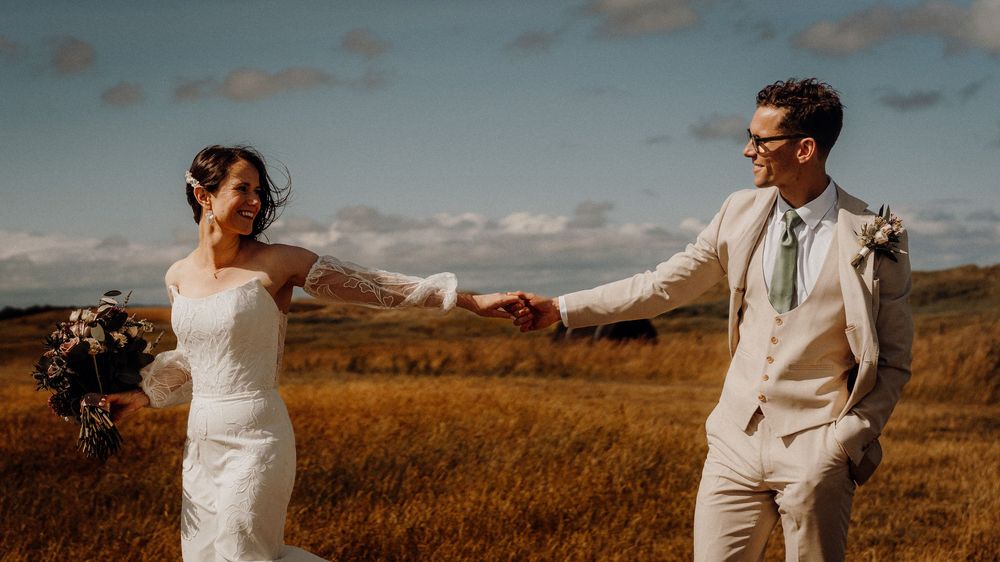 A couple in formal wedding attire embraces lovingly in an autumn field with golden grass at sunset.