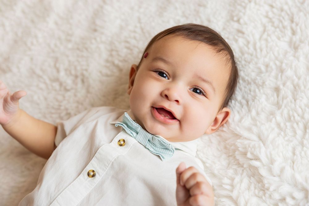Smiling baby wearing white outfit with blue bow tie lies on soft white textured blanket.