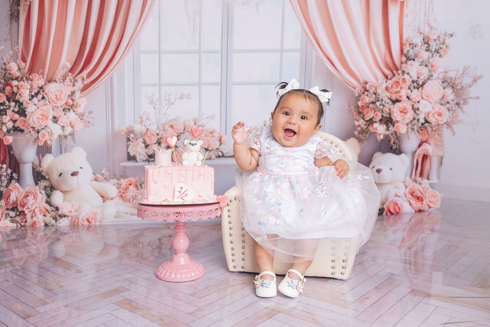 Pink and white first birthday party setup with floral decorations, cake, and baby shoes in a studio setting.
