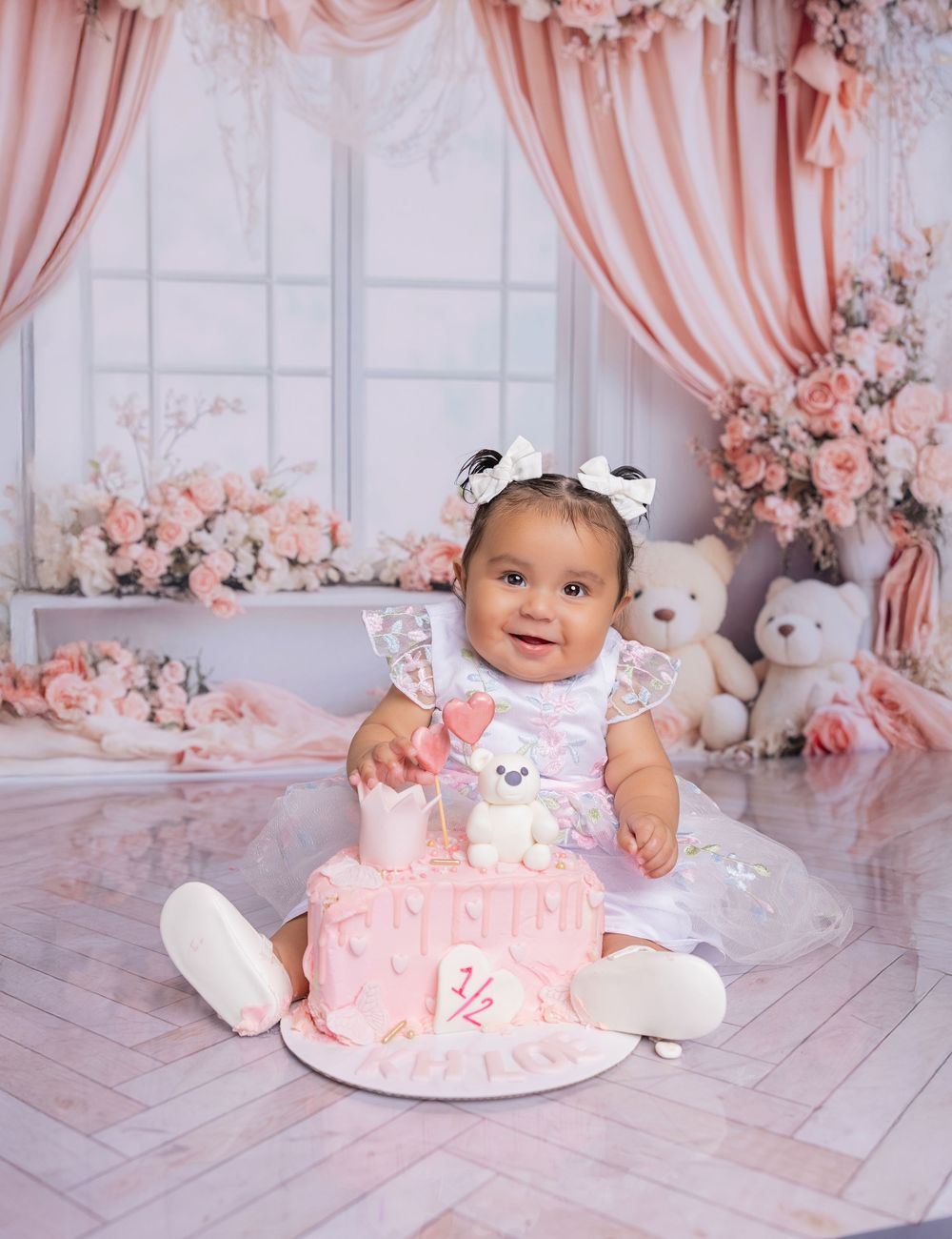 Baby in pink dress sits beside pink birthday cake with teddy bears and floral decorations in elegant studio setting.