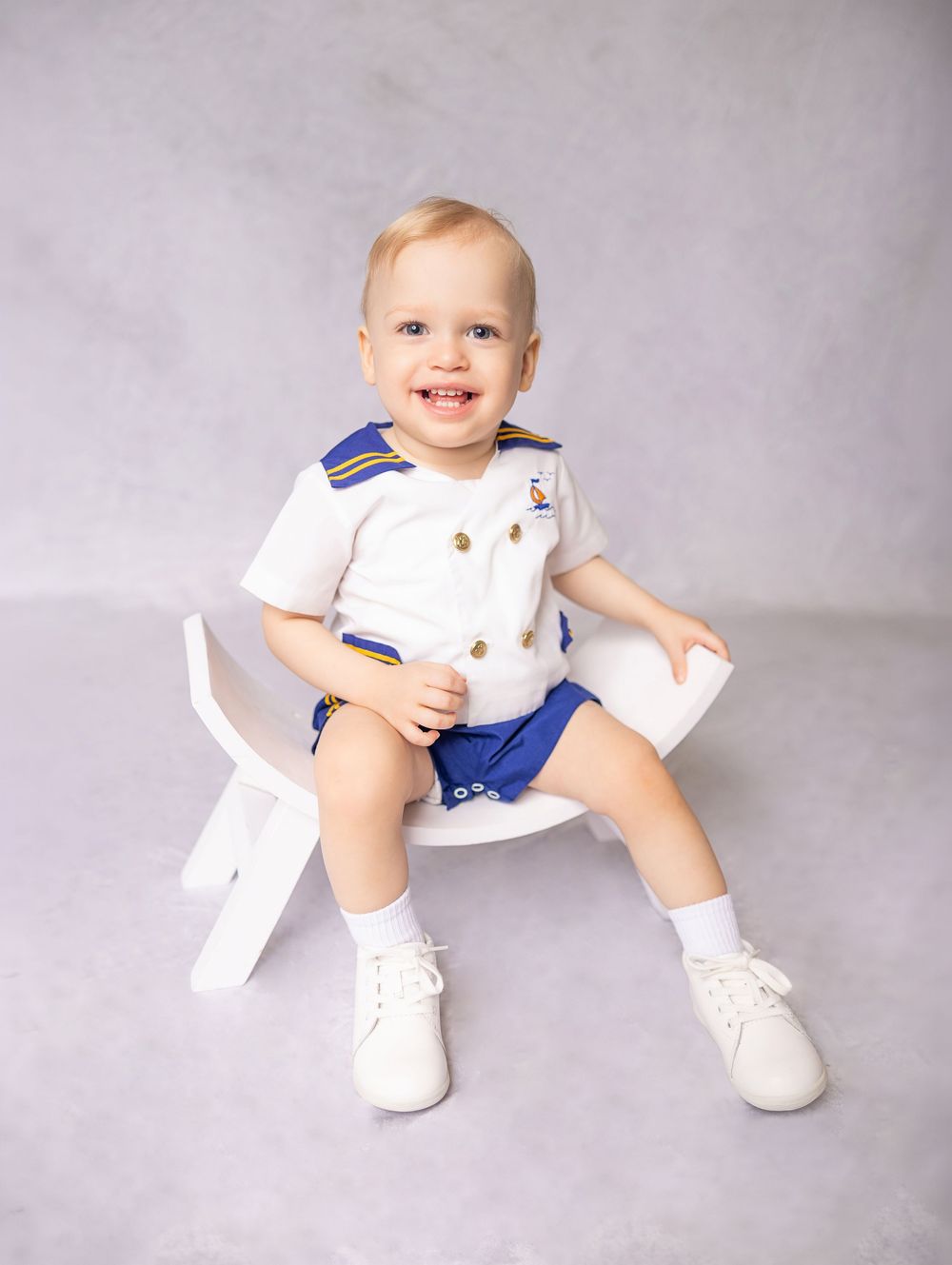 Smiling baby boy in white and blue sailor outfit sitting on white chair for a professional portrait photo.