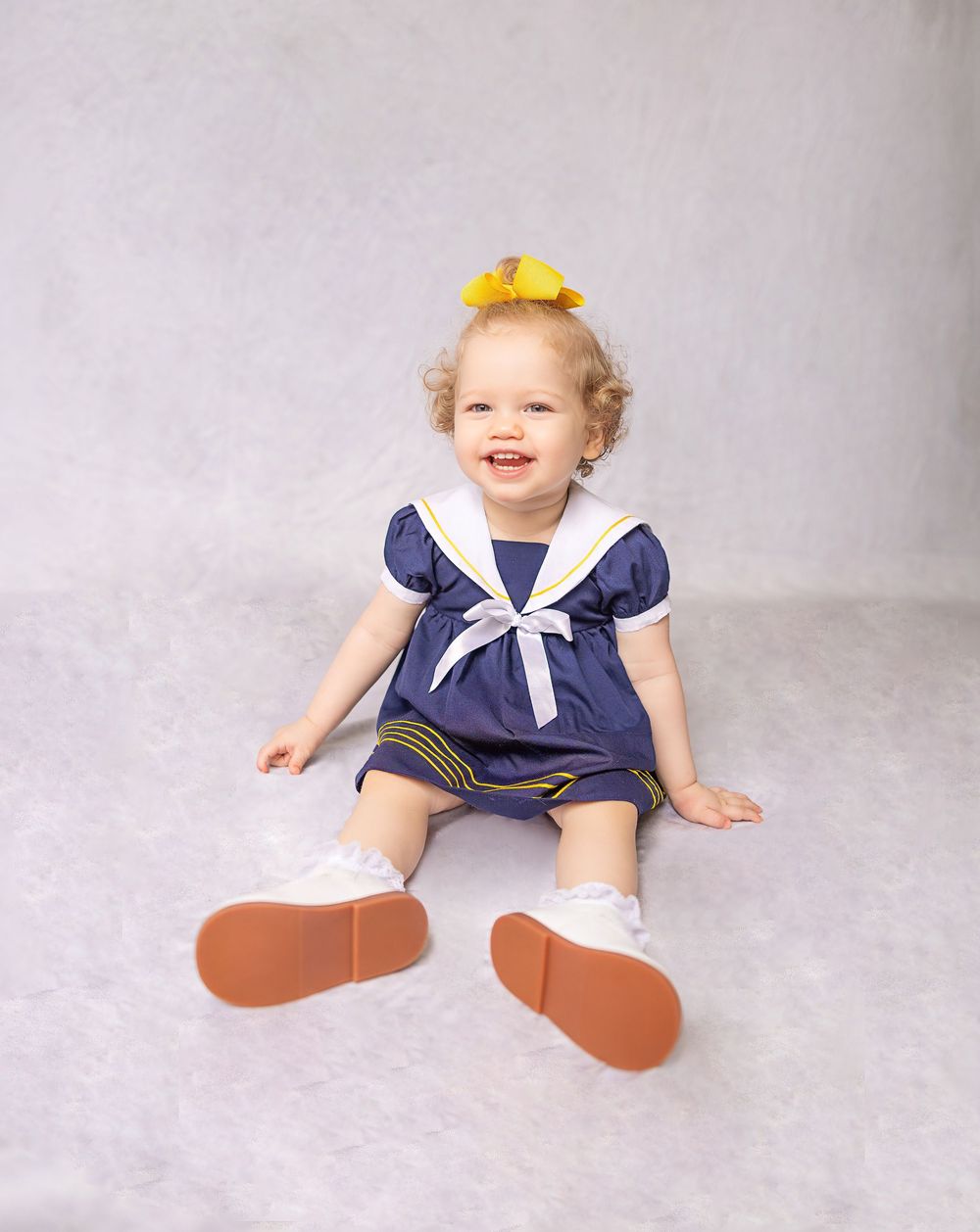 Toddler girl in navy sailor dress with yellow bow sits smiling on gray backdrop wearing white shoes.