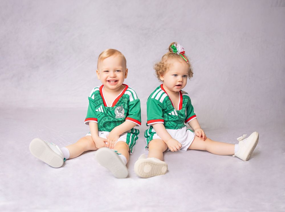 Two toddlers in matching green Adidas soccer jerseys sitting together for a studio portrait photo.