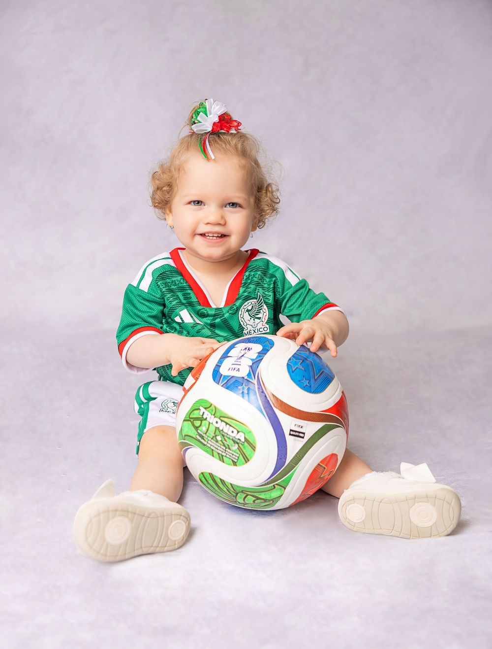 Smiling toddler girl in green Mexico soccer jersey holding a soccer ball, wearing white shoes and a colorful bow.